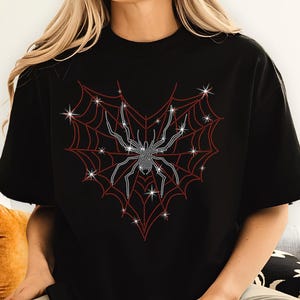 Pode incluir: Camiseta preta com um design de aranha e teia de strass. A aranha é prateada e a teia é vermelha.