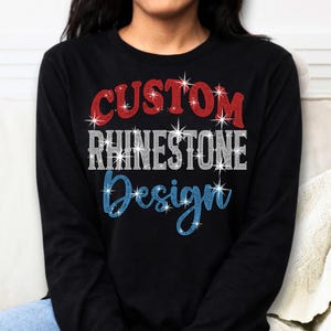 May include: Black long-sleeve shirt with the words "CUSTOM RHINESTONE Design" in red, white, and blue rhinestones. The shirt is a classic crew neck style and is made of a soft, comfortable material. The design is centered on the front of the shirt.