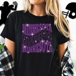 Könnte beinhalten: Schwarzes T-Shirt mit dem Wort "MINNESOTA" in einem lila, wellenförmigen Design mit Sternakzenten. Das Shirt wird mit einem schwarz-weiß karierten Hemd getragen. Eine Footballspieler-Silhouette und ein Helm-Grafik befinden sich im Hintergrund.