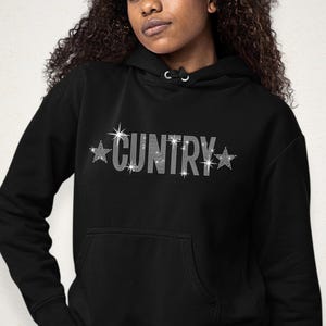 May include: Black hoodie with the word "CUNTRY" spelled out in sparkling rhinestones. The word is flanked by rhinestone starbursts. The hoodie has a front pocket and a hood with drawstrings.