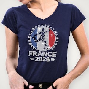 May include: Navy Bella Canvas v-neck t-shirt featuring a sparkling rhinestone “France 2026” soccer graphic. The design includes a soccer player silhouette over a French inspired flag background with royal blue, red, and silver rhinestone details and star accents around the border. The model is wearing a navy v-neck shirt.