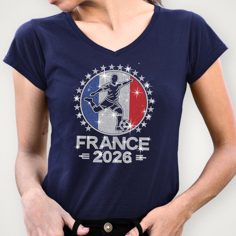 May include: Navy Bella Canvas v-neck t-shirt featuring a sparkling rhinestone “France 2026” soccer graphic. The design includes a soccer player silhouette over a French inspired flag background with royal blue, red, and silver rhinestone details and star accents around the border. The model is wearing a navy v-neck shirt.