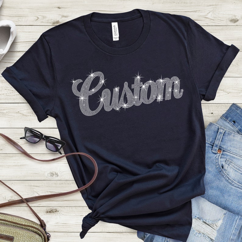 May include: Navy blue t-shirt with the word "Custom" spelled out in sparkling rhinestones. The shirt is a crew neck style and is knotted at the bottom. Sunglasses and a purse are visible in the background.