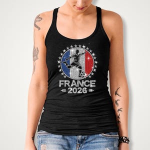May include: Black Bella Canvas tank top featuring a rhinestone “France 2026” soccer design. The artwork showcases a soccer player silhouette layered over a French inspired flag background with sparkling royal blue, red, and silver rhinestones surrounding the circular design. The model is wearing a black sleeveless tank top.