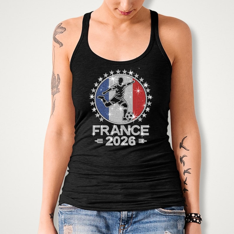 May include: Black Bella Canvas tank top featuring a rhinestone “France 2026” soccer design. The artwork showcases a soccer player silhouette layered over a French inspired flag background with sparkling royal blue, red, and silver rhinestones surrounding the circular design. The model is wearing a black sleeveless tank top.