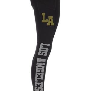 Può includere: Leggings neri con un design "LOS ANGELES" in strass bianchi sulla gamba sinistra e un design "LA" dorato sulla gamba destra.