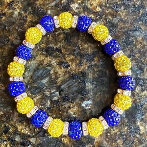 May include: A beaded bracelet featuring alternating yellow and blue crystal-covered beads. The bracelet is strung with a clear elastic cord and has small, rectangular silver-colored spacers between the beads. The bracelet is on a brown surface.