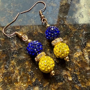 May include: A pair of gold-coloured earrings with blue and yellow beaded spheres. The earrings have a gold hook and are decorated with small, sparkling crystals. The earrings are displayed on a brown and gold speckled surface.
