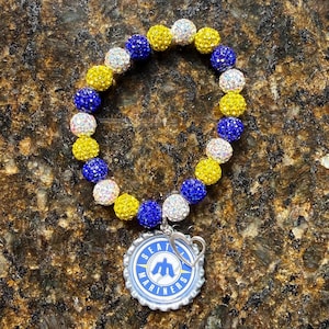 May include: A beaded bracelet featuring blue, yellow, and white sparkling beads. The bracelet is adorned with a Seattle Mariners bottle cap pendant. The pendant has a blue and white design with the team logo and text.