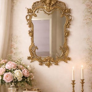 May include: An elaborate gold mirror with detailed carvings, hanging on a wall with floral wallpaper. A table below features a floral display, books, and candle holders, creating a classic, elegant scene.