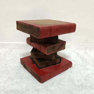 May include: A decorative side table crafted from stacked, faux books. The table features a red exterior with brown edges, resembling aged books. The design creates a unique, eye-catching piece for any room. The table measures approximately 20 inches tall.