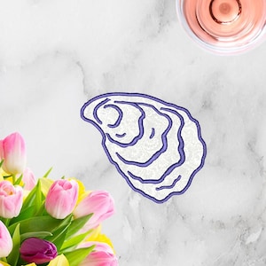 Op de afbeelding: Een geborduurd oesterschelpontwerp met een paarse omtrek en een wit interieur. De afbeelding bevat ook een boeket roze tulpen, gele narcissen en een glas roséwijn op een marmeren oppervlak.