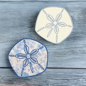 Puede incluir: Dos posavasos con forma de dólar de arena, uno con un patrón floral azul y el otro con un fondo crema. Ambos posavasos tienen un diseño de dólar de arena bordado en azul.