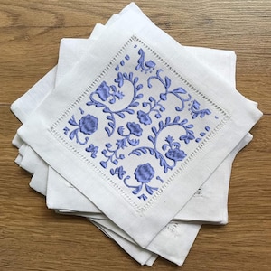 以下が含まれることがあります： 青い花の刺繍が施された白いリネン製のナプキンが積み重ねられています。ナプキンは折り畳まれ、木製の表面に置かれています。