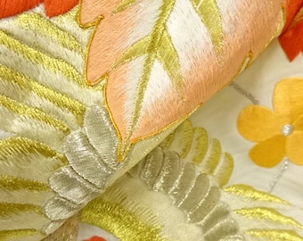 May include: Close-up of a white fabric with gold and silver embroidery. The embroidery features a floral design with leaves and flowers in shades of orange, yellow, and white.