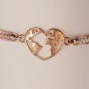 May include: A braided bracelet with a gold heart-shaped charm featuring a world map design. The bracelet is made with pink, white, and brown threads.