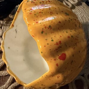 May include: A hollow, pumpkin-shaped ceramic vessel with a glossy orange exterior featuring speckled red and green paint splatters. The interior is smooth and white, with a large, irregular opening on one side and a ribbed, curved outer surface.