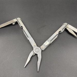 May include: A silver-colored multi-tool featuring a pair of pliers at the center hinge, with two folding handles that extend outward. One handle displays a partially deployed flat-head screwdriver, while the other shows a partially opened knife blade.