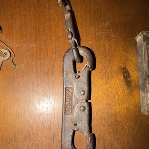 May include: A dark, weathered metal snap hook featuring a swivel top and a spring-loaded gate. The word COVERT is embossed vertically on the main body of the hook. The metal shows signs of oxidation and age, with a textured, dark brown finish.