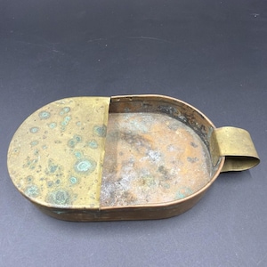 May include: An oval-shaped copper and brass container featuring a partial lid. The metal surfaces exhibit significant oxidation with green patina spots and weathered textures. A small, curved brass handle is attached to one side of the vessel.