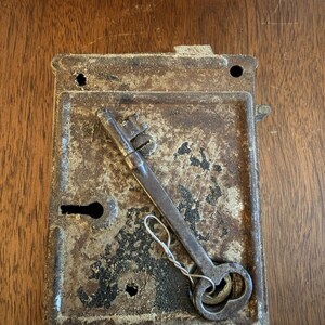 May include: A rectangular, weathered metal rim lock with a dark, rusted finish and visible surface wear. A long, slender metal skeleton key with a bow handle is positioned diagonally across the lock face, attached by a thin, twisted wire loop.
