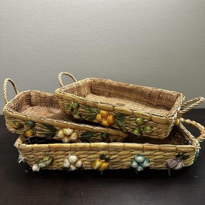 May include: Two rectangular woven baskets with handles, featuring decorative accents of yellow, white, blue, and purple garlic-shaped embellishments on the exterior sides. The baskets are stacked, showing a natural tan fiber texture throughout.