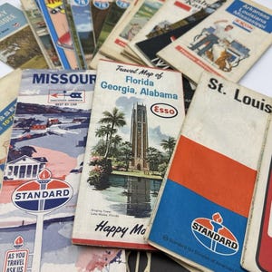 May include: A collection of vintage paper travel maps featuring various American states and cities. The covers display colorful illustrations of landmarks, oil company logos like Standard and Esso, and text including Missouri, Florida, and St. Louis.