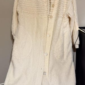 May include: A cream-coloured, button-down robe with long sleeves. The robe has a textured, ribbed design on the upper chest and a soft, plush texture throughout. Features two front pockets and a collar. The robe is hanging on a black hanger.