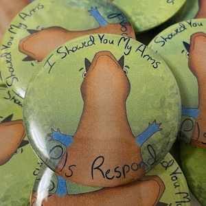 Puede incluir: Un botón verde con un lagarto de dibujos animados marrón con brazos azules. El texto en el botón dice "I showed you my arms, is respond".