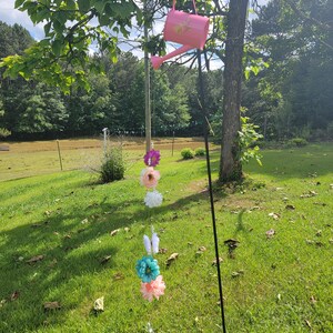 以下が含まれることがあります： 枝にピンクのジョウロがぶら下がっていて、そこから花と蝶の紐がぶら下がっています。花はピンク、白、青です。