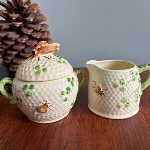 May include: A cream ceramic sugar bowl with lid and creamer set. The set has a textured surface, green shamrock and vine details, and a brown and orange insect design. The sugar bowl has a brown insect on the lid.