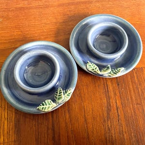 May include: Two blue ceramic egg cups with green leaf accents. The egg cups are designed to hold a single egg.