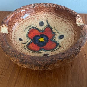 May include: A brown ceramic ashtray with a rough texture and a red, yellow and blue flower design.