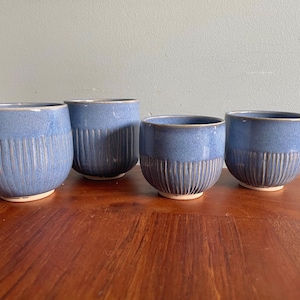 May include: Four blue ceramic bowls with vertical ribbed detailing on the lower half. The bowls have a rounded shape and a light-coloured base. The bowls are arranged on a wooden surface.