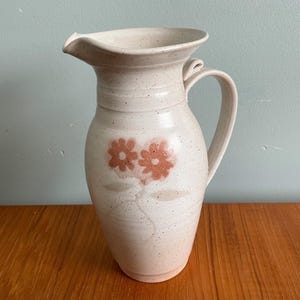 Peut inclure: Un pichet en céramique blanche avec un glaçage moucheté et un motif floral brun. Le pichet a une anse et un bec.