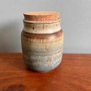 May include: A cylindrical ceramic storage jar with a cork lid. The jar features a speckled cream-coloured upper section that transitions into a warm brown and muted blue-grey glaze with vertical drip patterns throughout the body of the vessel.