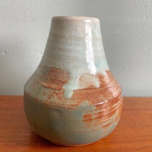 May include: A small, pear-shaped ceramic vase with a light blue glaze and brown accents. The vase has a slightly uneven surface and a rounded bottom.