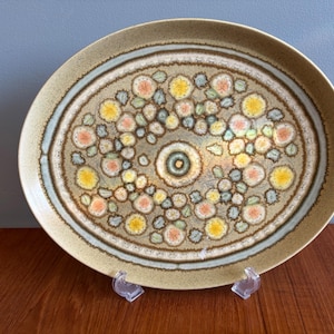 May include: Oval ceramic serving platter. The beige background features a central floral design in yellow, orange, and green. It has a light blue and brown border. The platter is displayed on a wooden surface.