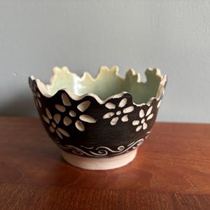 May include: A small ceramic bowl with a dark brown exterior featuring carved white flower patterns and a wavy, uneven rim. The interior of the bowl is glazed in a pale, light green colour, and the base is a plain, unglazed off-white ceramic.