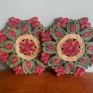 May include: Two floral coasters. Each coaster has a central red flower shape, encircled by green and red woven patterns. The centre is a natural beige colour. The coasters are for decorative use.
