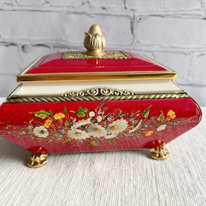 May include: A red and gold metal tin box with a floral design and a gold knob on the lid. The box has four gold feet.