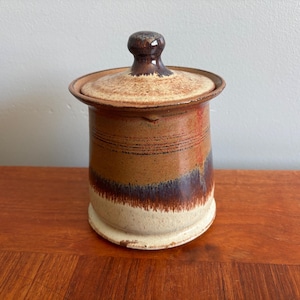 May include: A cylindrical ceramic canister with a matching lid featuring a knob handle. The exterior displays a multi-toned glaze with horizontal bands of tan, dark brown, and cream, accented by a subtle reddish-orange vertical streak.