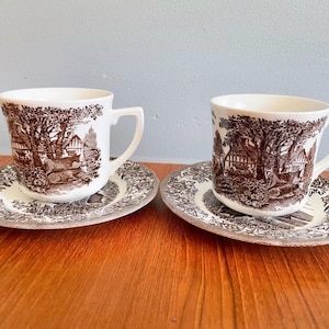 May include: Two brown and white teacups with matching saucers. The cups and saucers feature a brown and white floral pattern with a cottage scene.