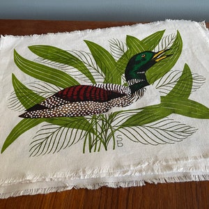 May include: A white linen placemat with a green and brown loon design. The loon is surrounded by green leaves and the placemat has a frayed edge.