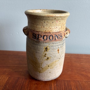 Peut inclure: Un pot en grès cylindrique avec un bord évasé et deux petites anses latérales. L'extérieur présente une partie supérieure beige mouchetée avec le mot SPOONS en relief en lettres sombres, et un corps inférieur gris et beige avec des motifs.
