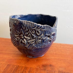 May include: A dark blue ceramic bowl with embossed floral and leaf patterns. The bowl has a textured surface and an uneven rim. The interior is a darker shade of blue, contrasting with the exterior. A handcrafted item.