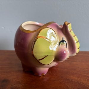 May include: A ceramic elephant-shaped planter or trinket holder featuring a glossy finish in shades of pink and yellow. The elephant has gold-painted trim along the ears, trunk, and top rim, with painted black eyes and eyelashes on its face.
