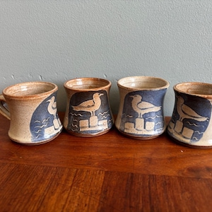 Peut inclure: Quatre tasses en céramique faites à la main sur le thème côtier. Chaque tasse présente un motif beige et bleu avec un goéland perché sur une jetée. Les tasses ont un aspect rustique et texturé, avec un bord et une anse bruns.