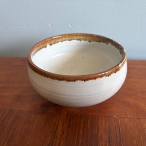 May include: A small, round ceramic bowl with a smooth, off-white interior and exterior glaze. The rim features a distinct, textured brown drip pattern that contrasts with the lighter body of the bowl, which has subtle horizontal ridges.
