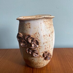 May include: A speckled ceramic vase with a light beige base and brown accents. The vase features a textured surface and a decorative cluster of brown leaves. The vase is displayed on a wooden surface.